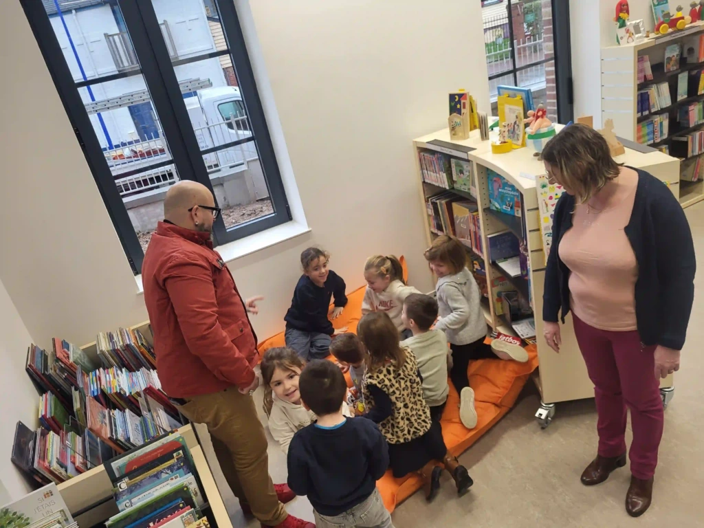 Enfants réunis dans l’espace jeunesse de la médiathèque du Crotoy, participant à une activité de lecture accompagnés par deux adultes, au milieu des rayonnages de livres.
